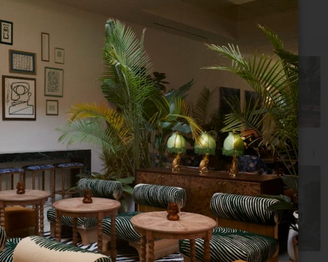 A stylish interior space features green plants, art-covered walls, and seating with striped upholstery paired with wooden tables.