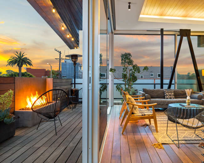 A contemporary outdoor living space features a modern fire pit, stylish seating arrangements, and large glass windows with a sunset view.