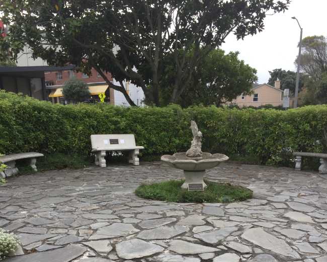 The image shows a circular stone courtyard with a fountain at its center, surrounded by benches and greenery.