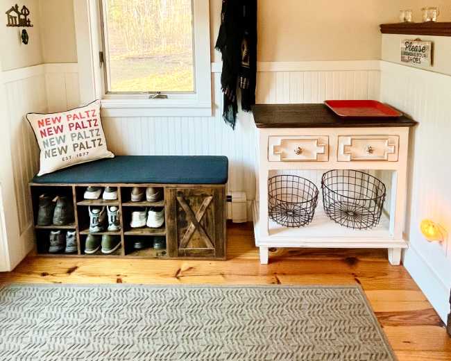 The image shows a cozy entryway featuring a shoe storage bench, a wall-mounted coat rack, a small table with decorative baskets, and a window with natural light illuminating the space.