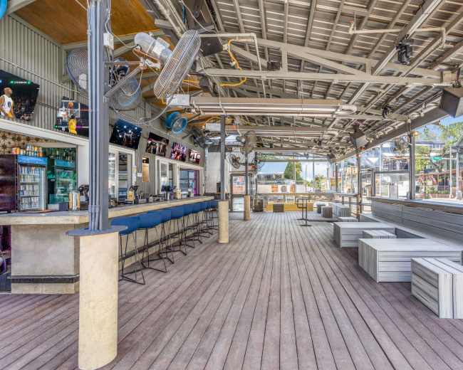The image shows a spacious, partially covered bar area with wooden flooring, blue stools along the counter, and several seating units arranged in the open space.