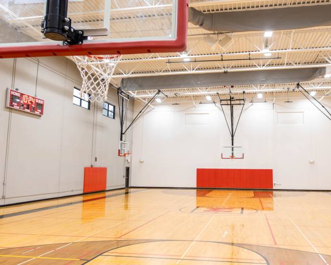 The image shows a spacious indoor basketball court with two hoops, a scoreboard, and polished wooden flooring.