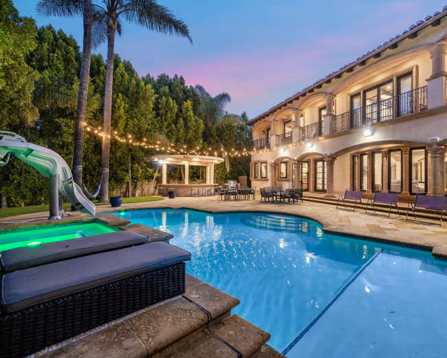 A luxurious backyard features a large swimming pool with a water slide, surrounded by palm trees and a two-story house with outdoor seating and string lights.