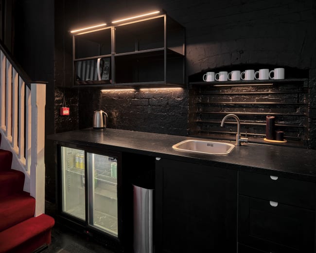 The image shows a modern kitchen area with dark cabinetry, a sink, a kettle, and an under-counter fridge, all lit by recessed lighting.