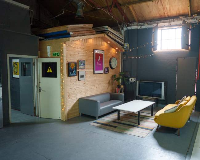 The image shows a spacious room with a gray couch, a yellow chair, a wooden wall, a television, and a window letting in natural light, adorned with string lights.