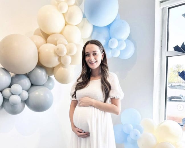 A woman in a white dress stands in front of a wall decorated with clusters of blue and beige balloons, holding her pregnant belly.