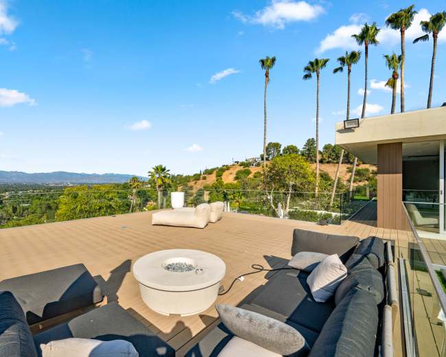 The image shows a spacious outdoor deck with modern seating and a fire pit, surrounded by palm trees and a scenic view of rolling hills and mountains.