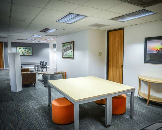 The image shows an office space with a large table, round orange stools, and a seating area, featuring various artworks on the walls.
