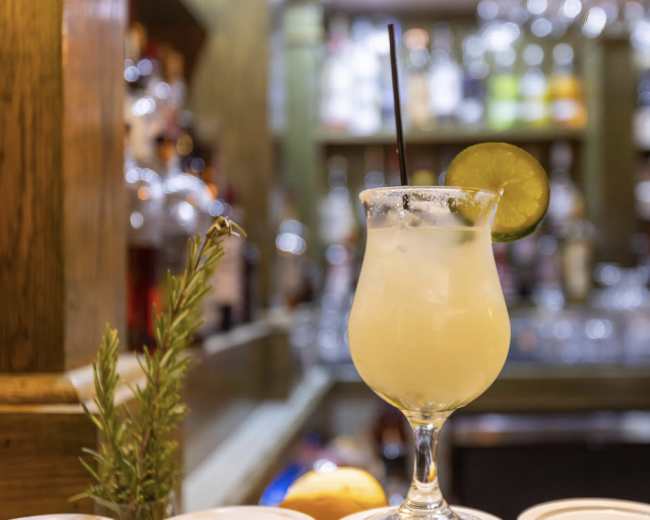 A cocktail with a lime slice and a straw sits on a bar counter with bottles in the background.
