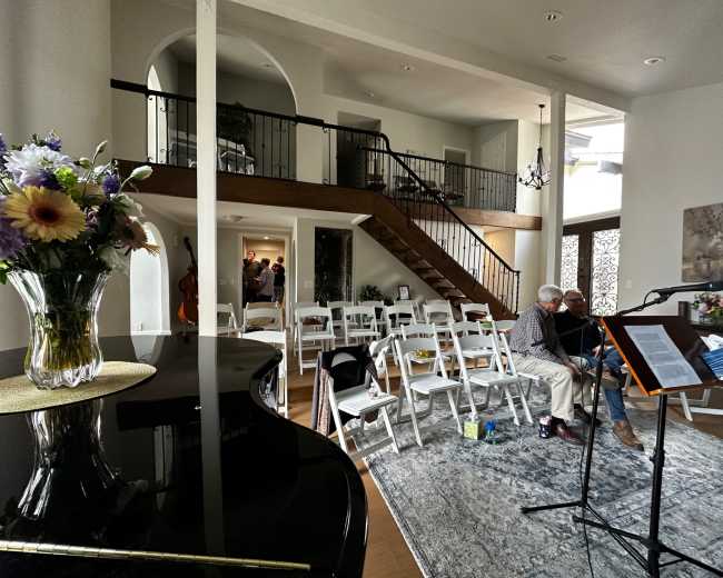 A spacious living area features a grand piano, a staircase, and rows of white chairs set up for an event, with individuals conversing in the background.
