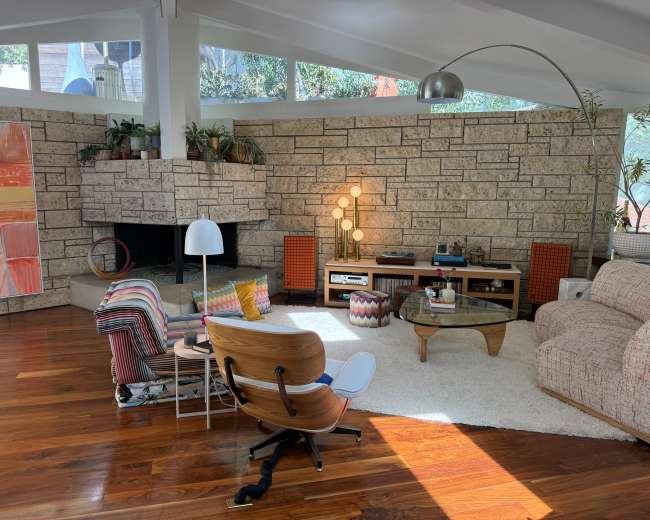The image shows a mid-century modern living room featuring a stone fireplace, a large window allowing natural light, and various seating options arranged around a circular coffee table.