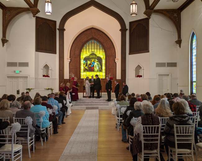 A wedding ceremony is taking place in a church, with a group of people gathered at the front and attendees seated in rows facing them.