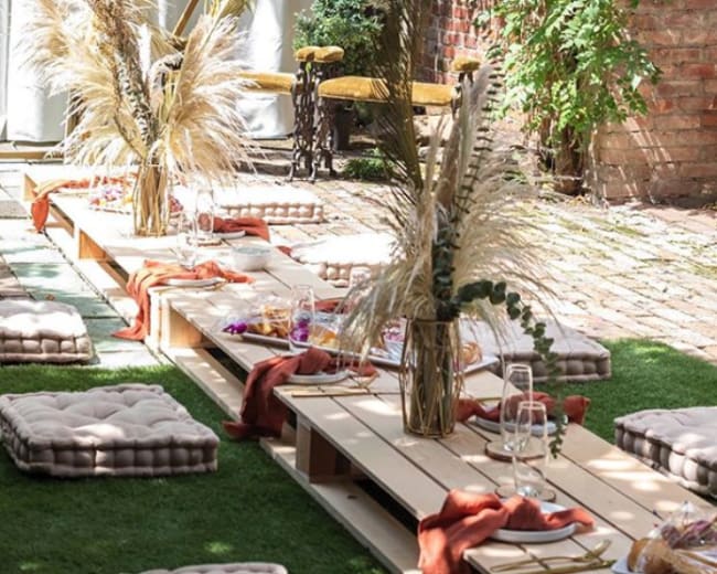 A decorated outdoor space with a wooden platform adorned with cushions and floral arrangements, surrounded by greenery and a backdrop of a white curtain.