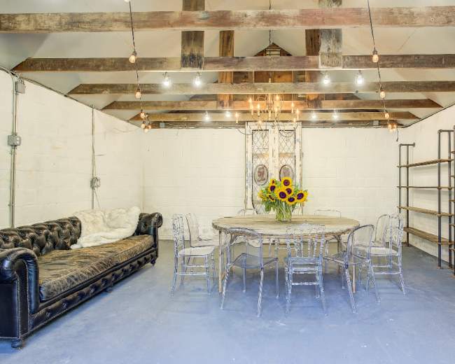 A spacious room features a round dining table surrounded by chairs, a black leather couch, and open shelves against the wall, with exposed beams and pendant lighting above.