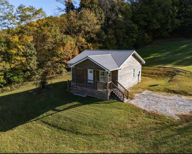 A small wooden cabin with a metal roof sits on a grassy area surrounded by trees.