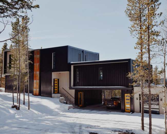 A modern two-story house with a mix of dark and light exterior materials is situated among snow-covered trees.