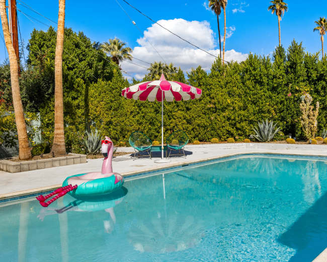 A pink flamingo pool float rests in a clear swimming pool surrounded by palm trees and lush greenery under a blue sky.