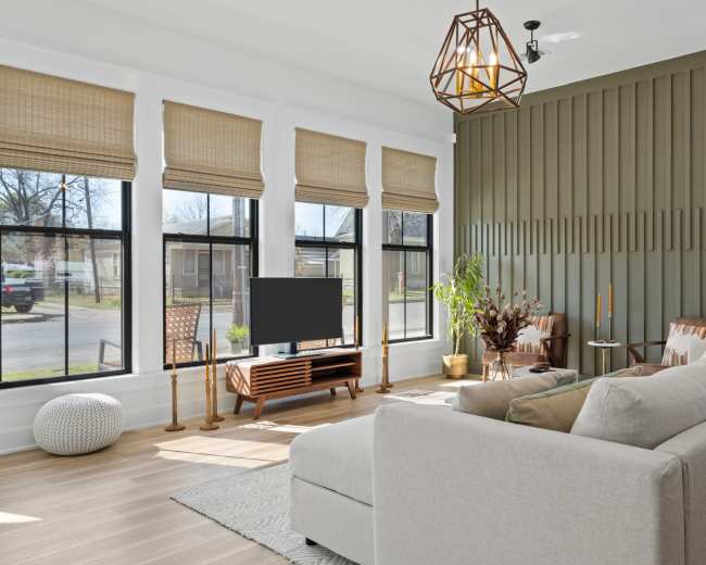 A modern living room features a sectional sofa, a wooden TV stand with a television, and large windows adorned with woven shades, all set against a green accent wall.