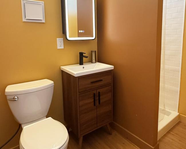 The image shows a small bathroom featuring a toilet, a wooden vanity with a sink, a mirror, and a shower stall, all set against yellow walls.