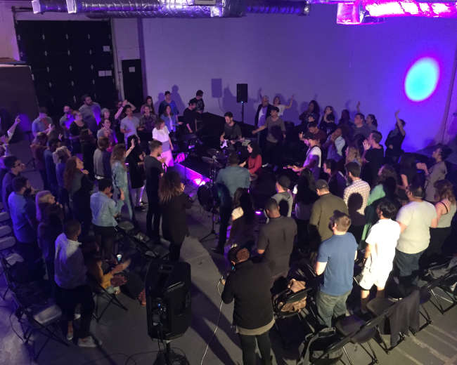 A group of people stands in a dimly lit space, gathered around musicians performing on a small stage.