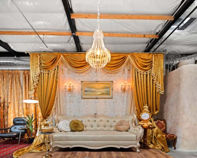 The image shows a elegantly decorated interior space featuring a light-colored sofa, ornate curtains, a chandelier, and a plush white rug on a wooden floor.