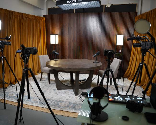 The image shows a recording studio setup featuring a round table surrounded by two chairs, with several cameras on tripods and lighting equipment in the background.