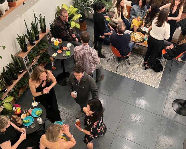 A group of people socializing at a gathering in a room filled with plants and small tables, with food and drinks on display.