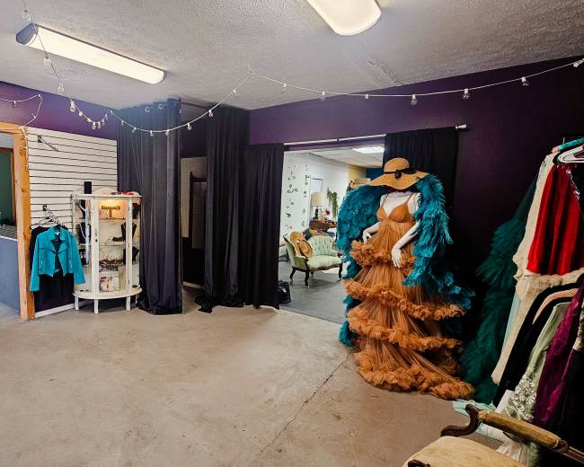 The image shows a room with a purple wall, featuring mannequins dressed in colorful outfits, a display case, and a doorway leading to another space.