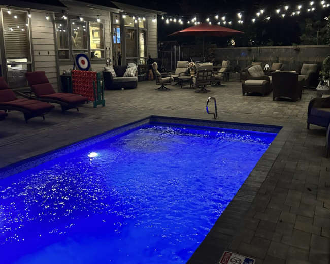 The image shows a well-lit backyard with a rectangular swimming pool illuminated in blue, surrounded by patio furniture and string lights overhead.