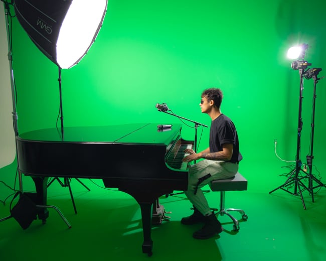 A person sits at a grand piano in front of a green screen, with studio lights positioned nearby.