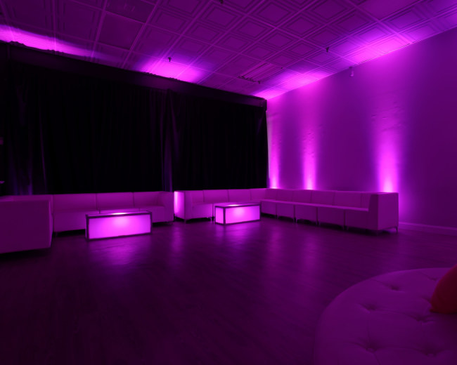 A modern lounge space with purple lighting, featuring white seating and illuminated coffee tables.