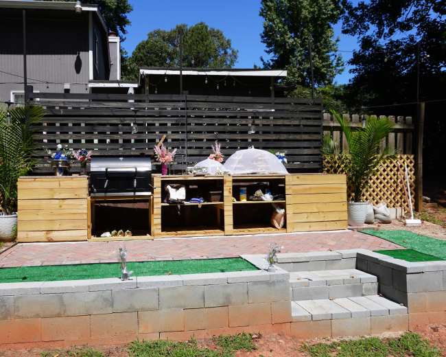The image shows an outdoor kitchen setup with a wooden counter, grill, and storage shelves, surrounded by a landscaped area with pavers and plants.