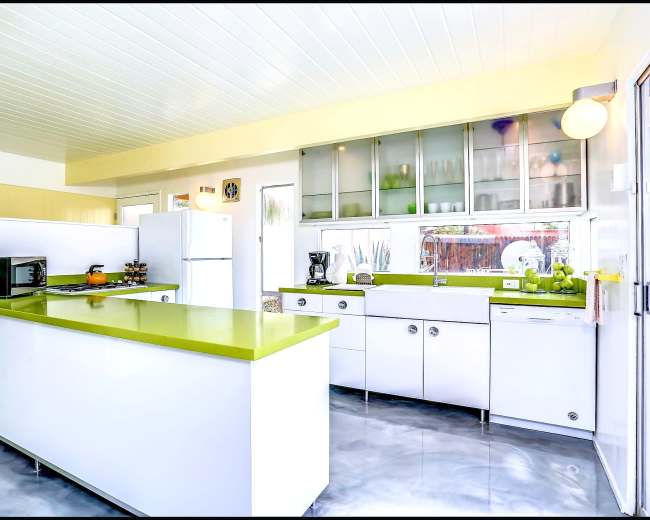 The image shows a modern kitchen with white cabinetry and green countertops, featuring a refrigerator, microwave, and coffee maker on a large island.