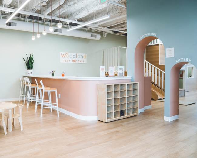 The image shows a brightly lit play care facility with a reception area, children’s seating, and a staircase leading to a play area.