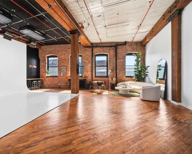 The image shows a spacious photo studio featuring wooden floors, exposed brick walls, and a large white backdrop, with a sectional couch and plants positioned in the corner.