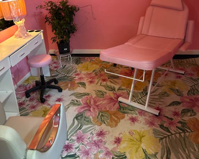 The image shows a small, brightly colored room with pink walls, a pink treatment chair, a white desk with a lamp, and a floral rug.