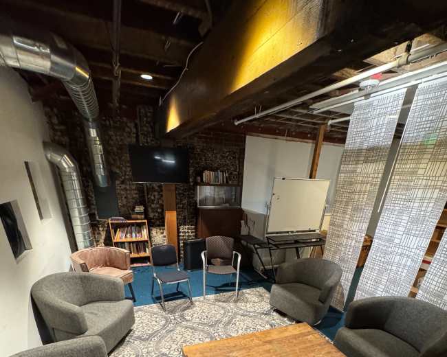 The image shows a communal seating area with several chairs arranged around a wooden coffee table on a patterned rug, set against a backdrop of exposed brick walls and wooden beams.
