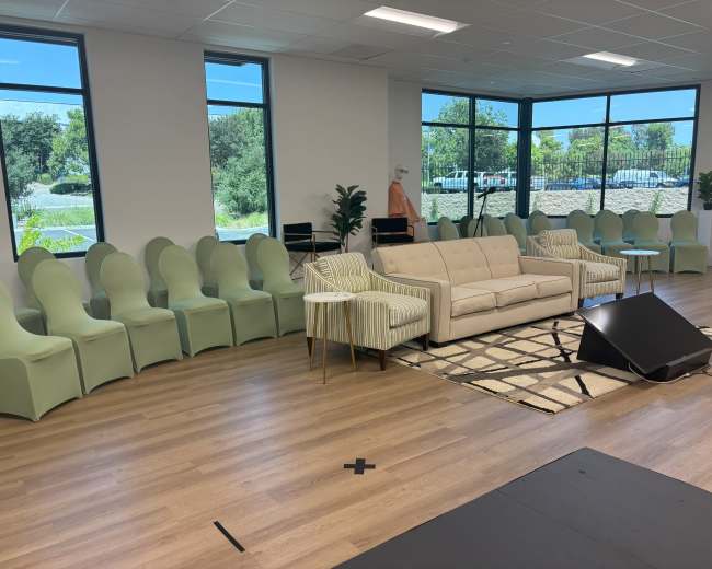 The image shows a modern room with a light-colored couch and striped armchair facing a row of green chairs against a wall of large windows.