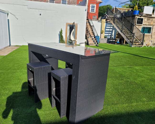 A modern outdoor bar table with two stools is set on artificial grass in a yard, with steps leading to a building in the background.