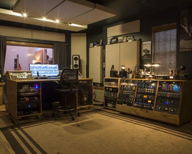 The image shows a music recording studio featuring a central mixing console, various audio equipment, and acoustic panels.