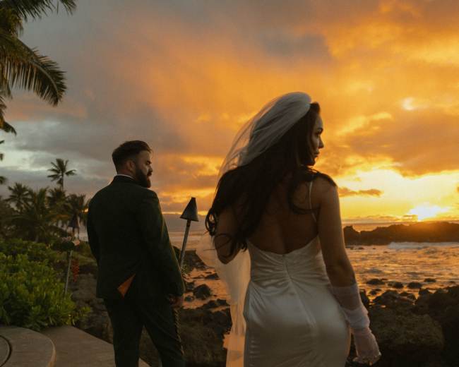 A couple stands by the ocean at sunset, with palm trees in the background and waves crashing on the rocks.