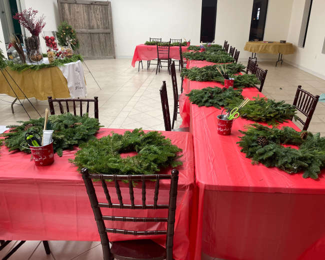 A spacious indoor setting with several red-covered tables arranged in rows, each adorned with green wreaths and decorative supplies in red containers.