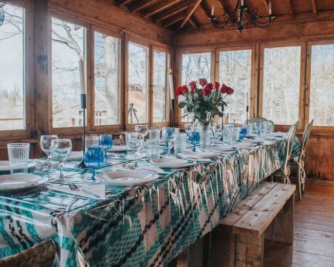 A long dining table is set with glassware and plates, adorned with a patterned tablecloth, in a room surrounded by large windows.