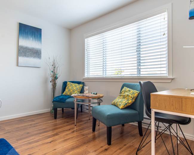The image shows a well-lit room featuring two teal chairs with decorative cushions, a small coffee table between them, a wooden desk with a plant, and a large window with blinds.