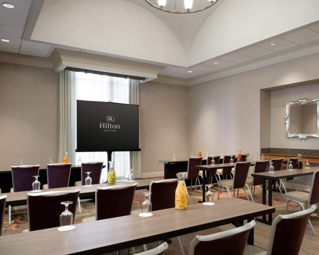 The image shows a conference room setup in a Hilton hotel, featuring tables with chairs arranged for a meeting, a large screen displaying the Hilton logo, and refreshments on the side.
