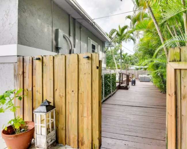 A wooden fence opens to a narrow walkway leading to a deck surrounded by tropical plants.