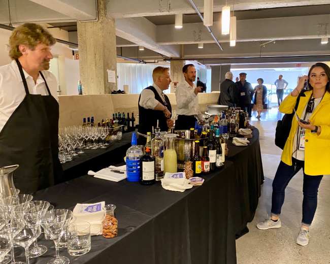 A group of people stands behind a bar laden with various bottles and glassware, while a woman dressed in a yellow coat converses on her phone nearby.