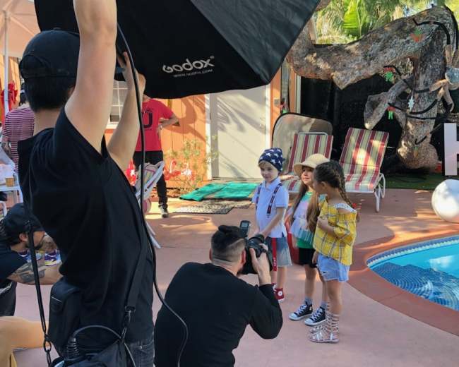 A photographer captures a group of children posing together in a backyard setting with a swimming pool and lounge chairs.