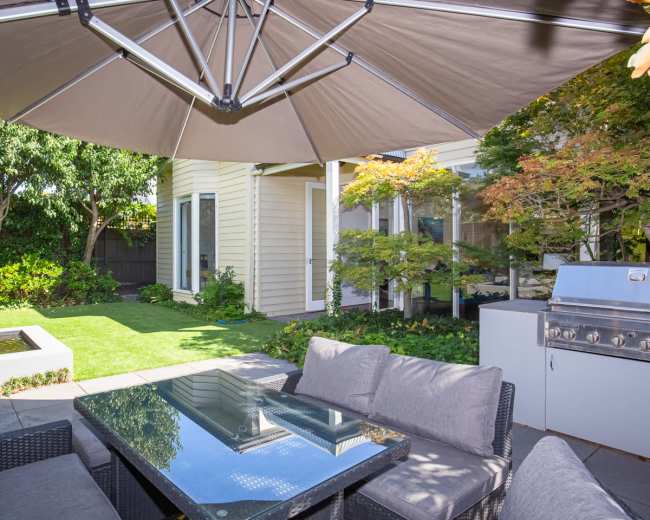 A patio area features a glass-top table surrounded by cushioned seating, with an umbrella above and a grill nearby, adjacent to a landscaped garden and a house.