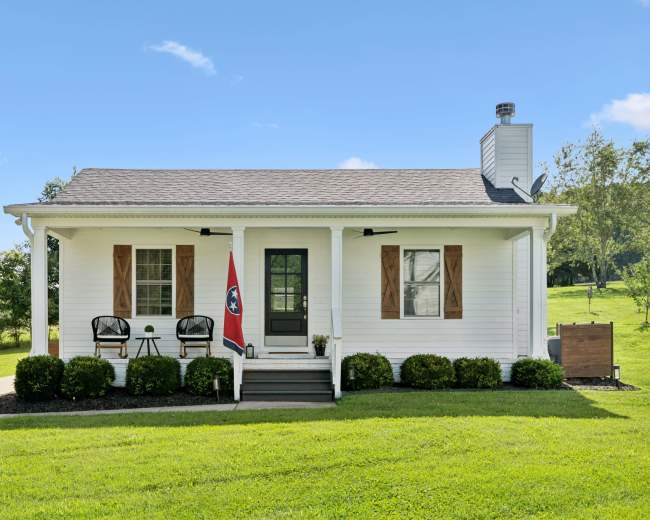 The image shows a single-story white house with a brown roof, featuring a small porch with two black chairs and a Tennessee flag.
